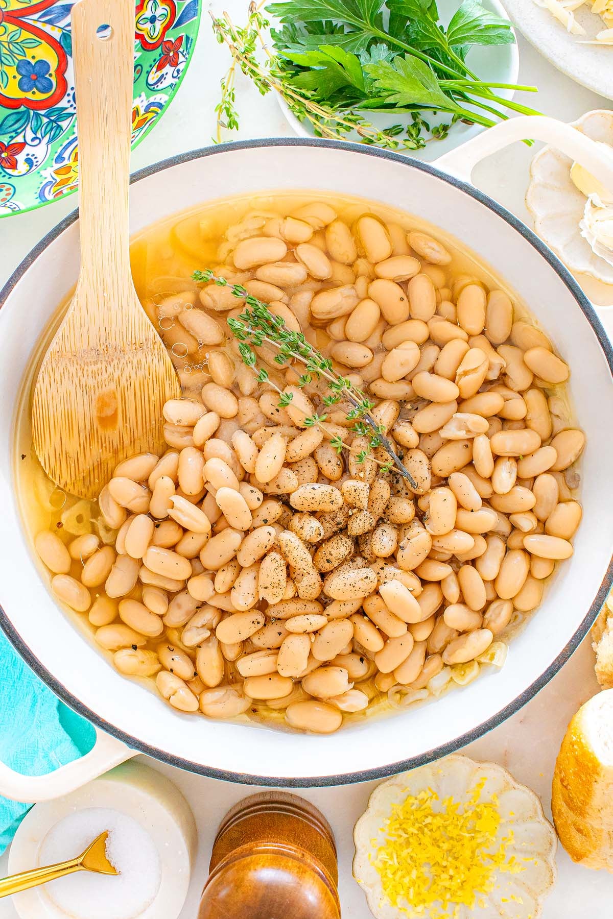 Tuscan white beans in broth with herbs and pepper simmer in a pot, a wooden spoon resting inside, surrounded by fresh ingredients.