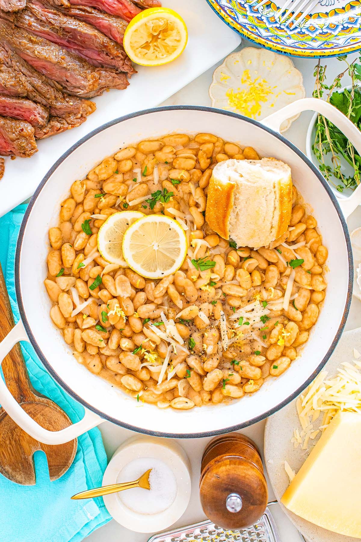 Tuscan white beans in a skillet, topped with lemon slices, herbs, cheese, and bread. Sliced steak is nearby.