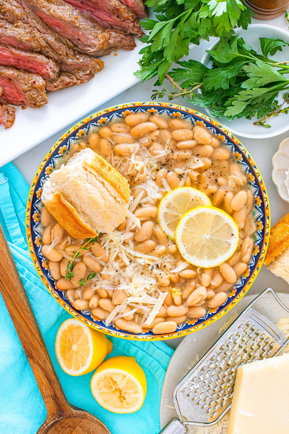 Bowl of Tuscan white bean soup with lemon slices, grated cheese, bread, parsley, and sliced steak on the side.