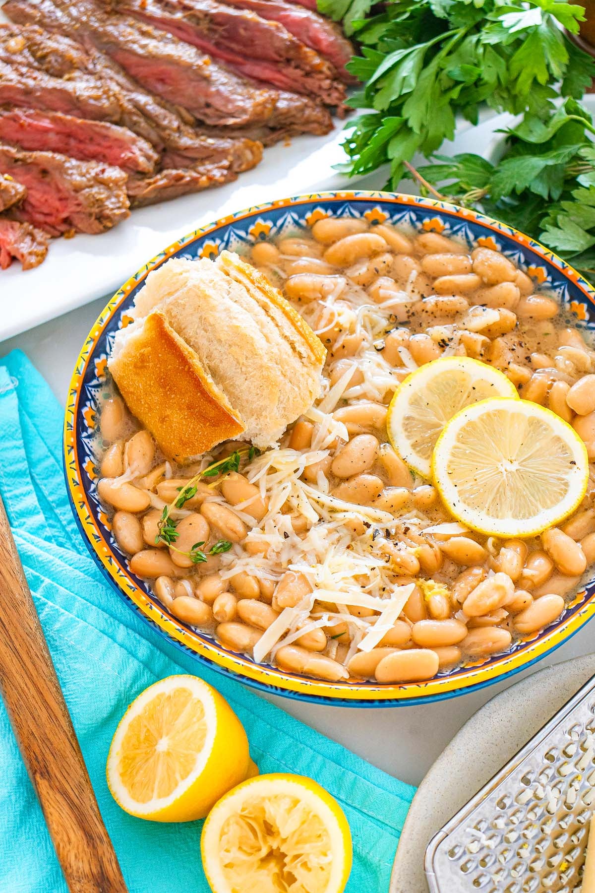A bowl of Tuscan white bean soup with lemon slices, bread, grated cheese, herbs, and sliced steak on the side.