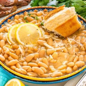 A bowl of Tuscan white bean soup with lemon slices and bread, surrounded by herbs and sliced steak.