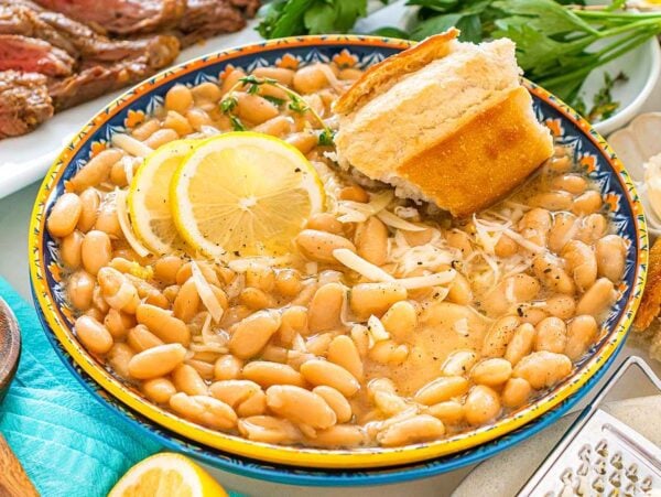 A bowl of Tuscan white bean soup with lemon slices and bread, surrounded by herbs and sliced steak.