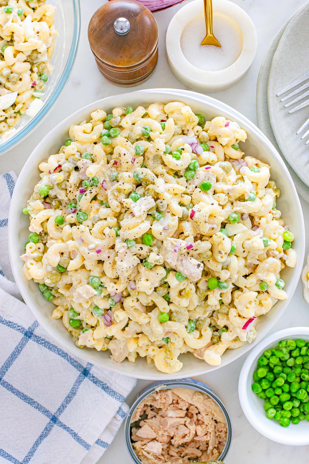 A bowl of creamy tuna macaroni salad with peas, tuna, and red onions, surrounded by ingredients and a napkin.