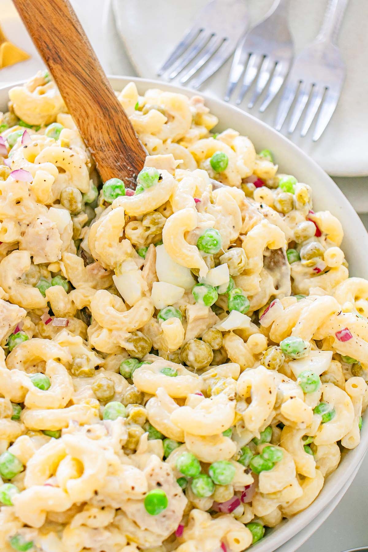 Close-up of tuna macaroni salad with peas, eggs, and red onions in a bowl with a wooden spoon.