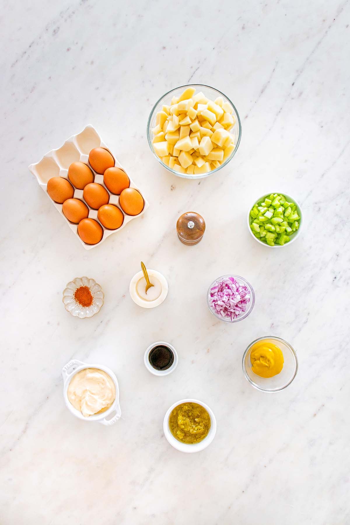 Ingredients for potato salad, including eggs, potatoes, celery, onion, mustard, mayonnaise, and spices on a white surface.
