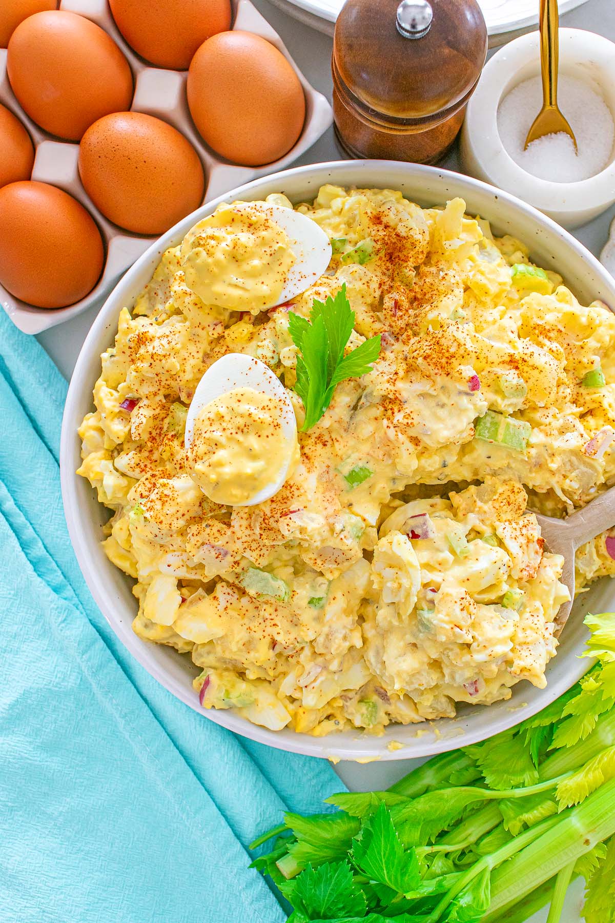 Bowl of creamy egg salad with sliced eggs, celery, and paprika, surrounded by whole eggs and seasonings.