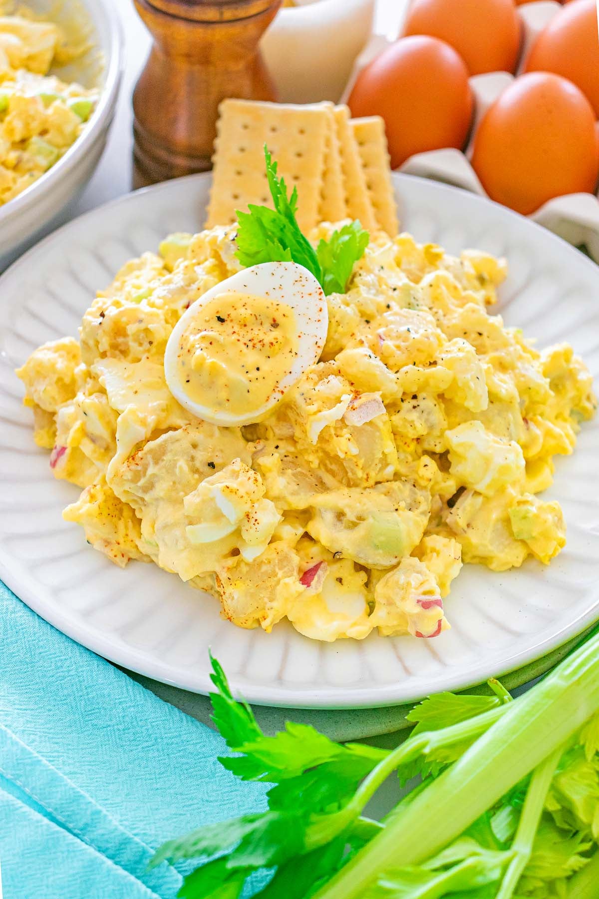 Potato salad topped with a halved boiled egg, crackers, and parsley, with celery and eggs in the background.