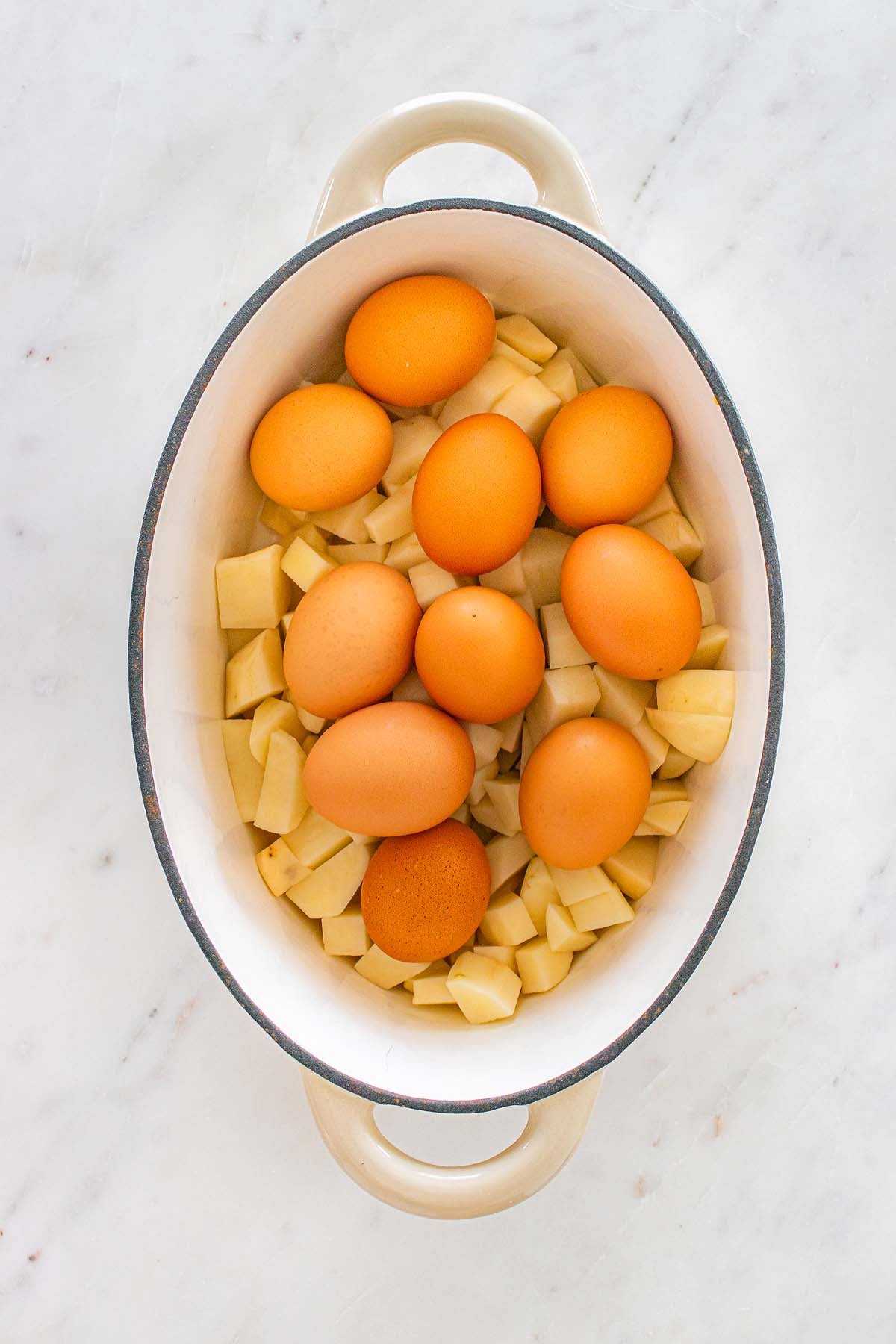 Brown eggs on top of chopped potatoes in a white oval casserole dish on a marble surface.