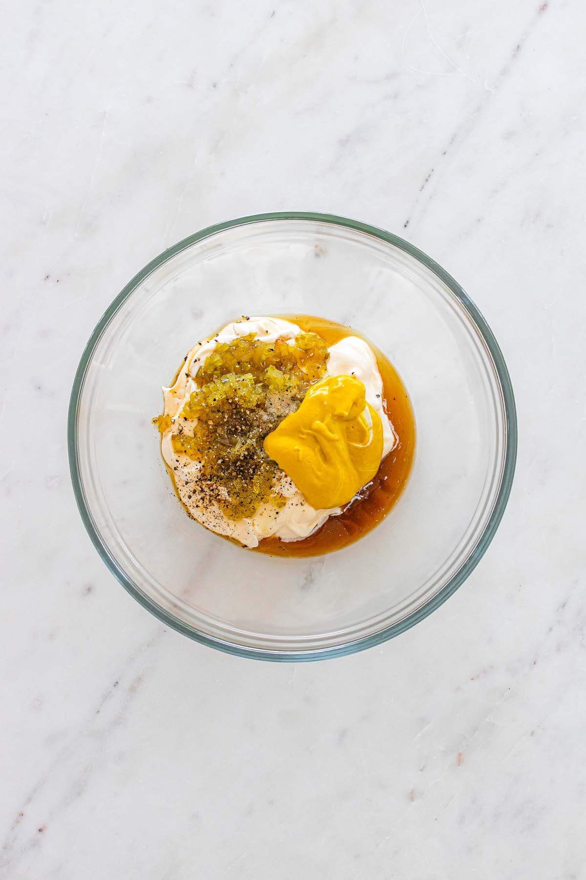 Glass bowl with mayonnaise, mustard, relish, and spices on a white marble surface.