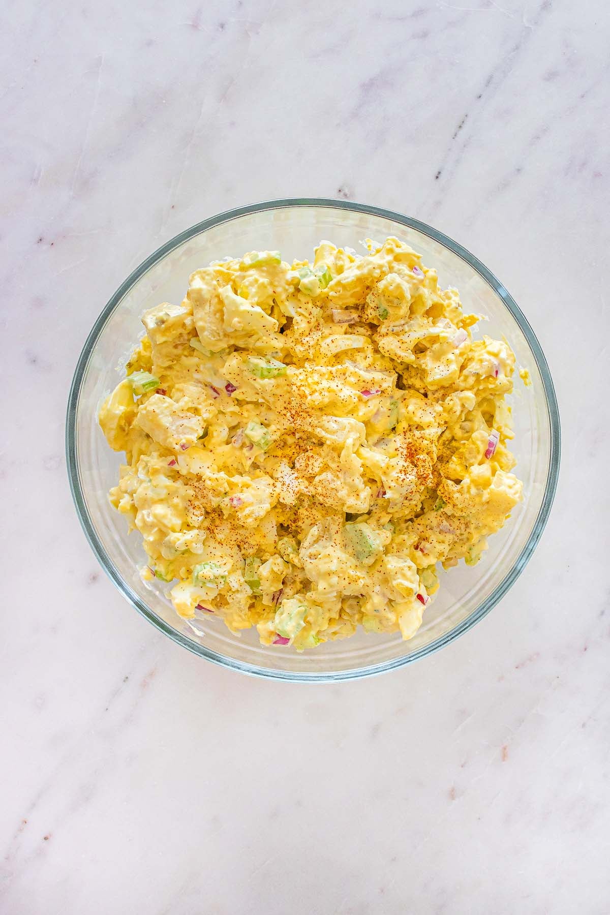 A glass bowl filled with creamy potato salad, garnished with paprika, on a white marble surface.