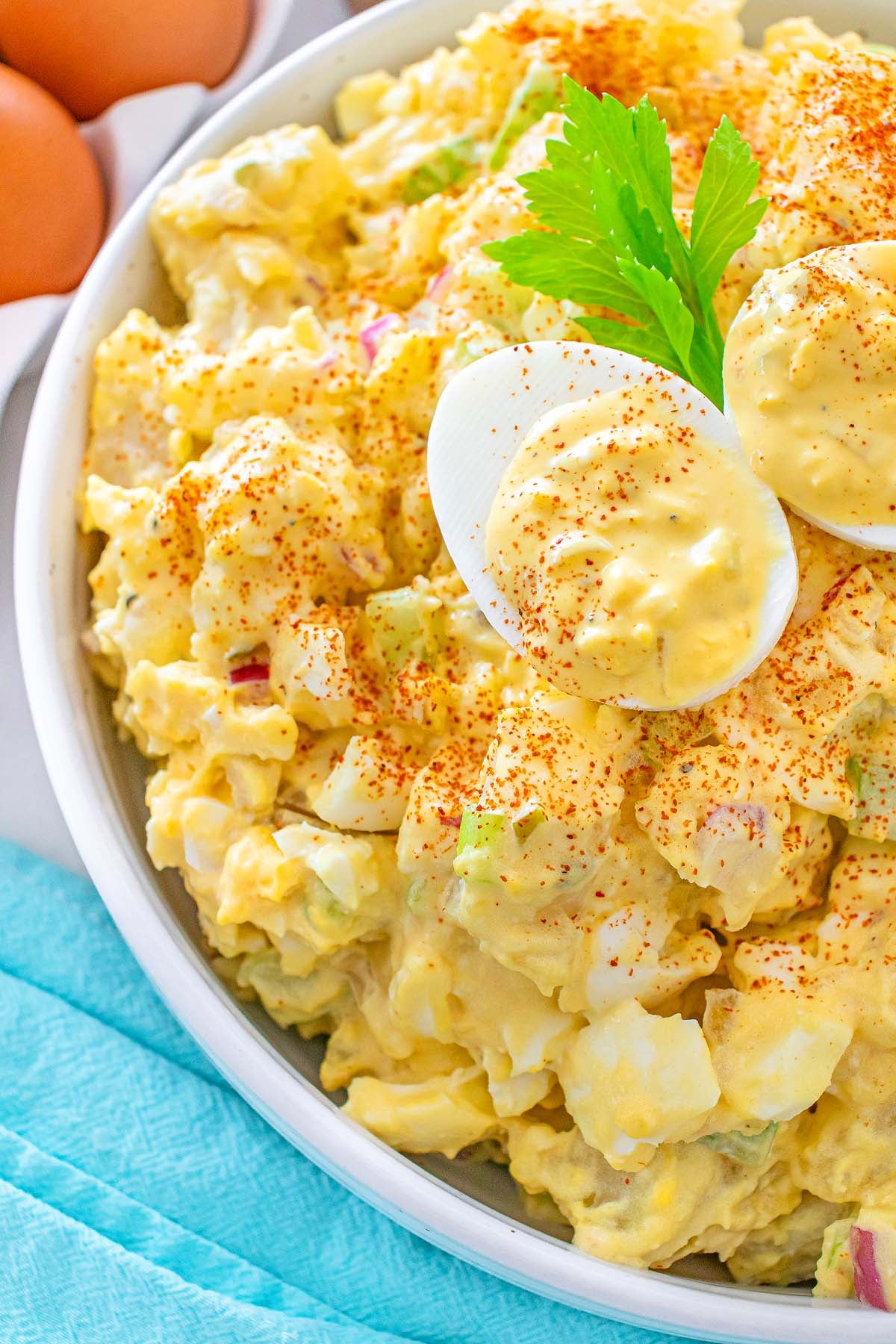 Bowl of creamy potato salad topped with sliced hard-boiled eggs, paprika, and a sprig of celery.