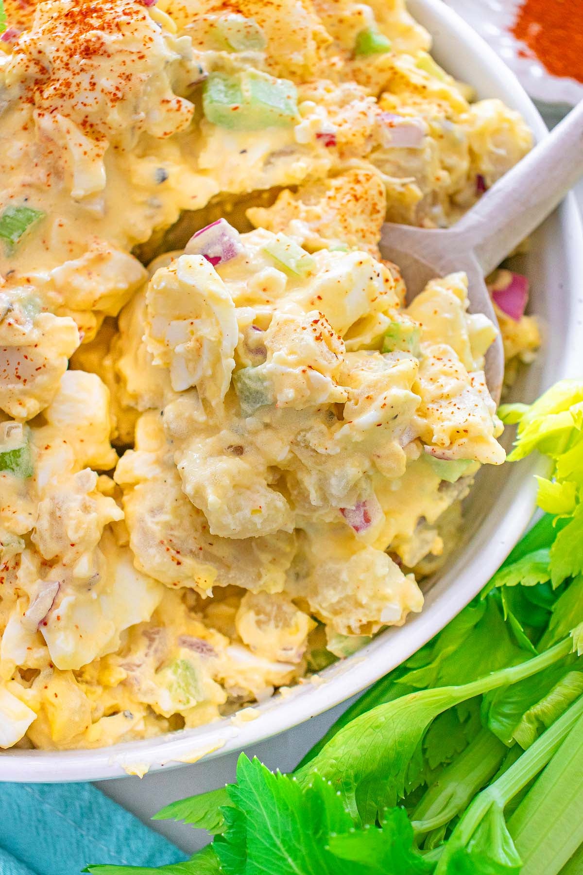 Creamy potato salad with chopped eggs, celery, and seasoning in a white bowl, next to fresh celery stalks.