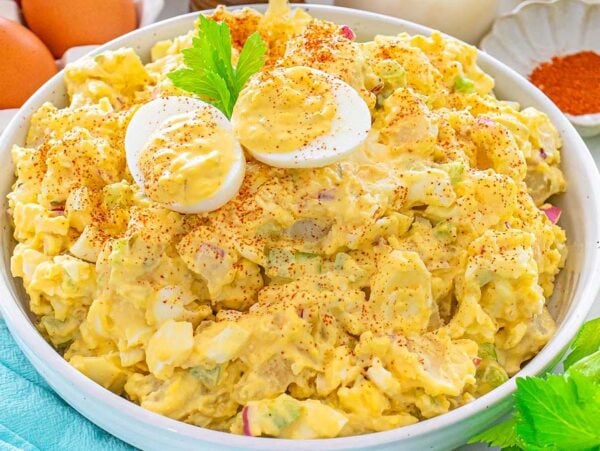 Bowl of potato salad topped with sliced boiled eggs and paprika, with eggs and celery nearby.