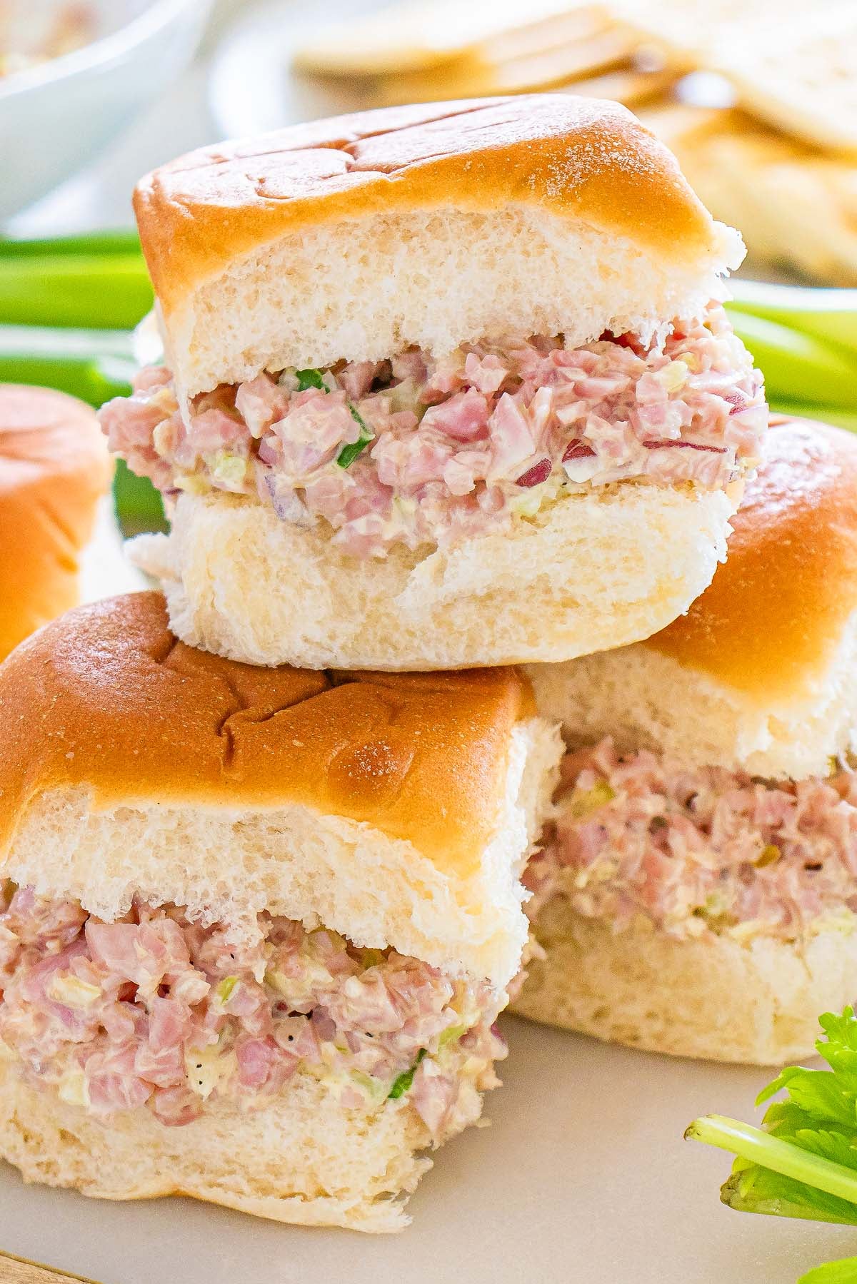 Three ham salad sliders with creamy filling on soft buns, stacked on a white surface with crisp celery in the background.