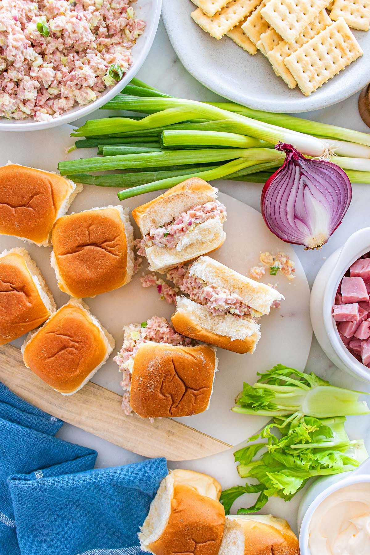 Enjoy delicious ham salad sliders on soft rolls, served with buttery crackers, crisp celery, green onions, and red onion mixed with savory ham chunks—all arranged beside a vibrant blue napkin.