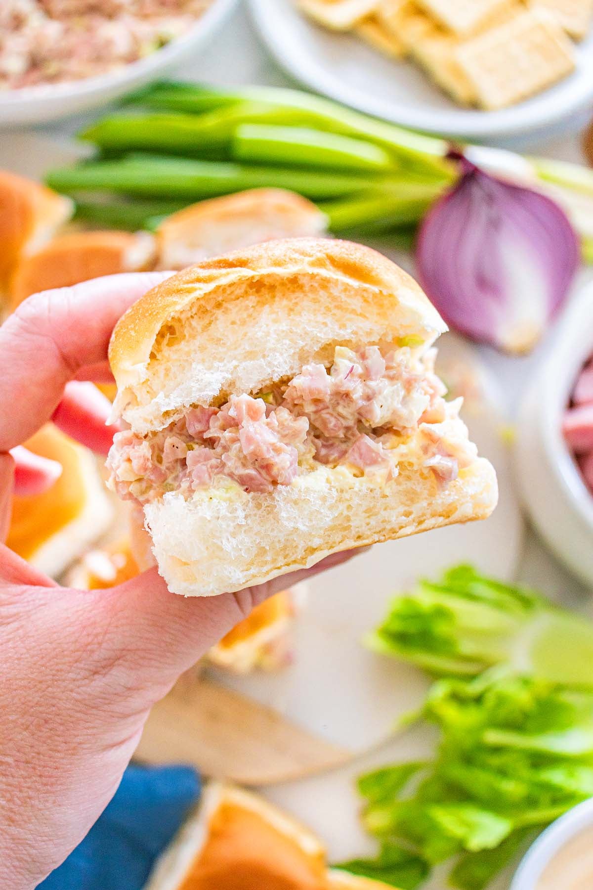 A hand holds a delicious ham salad sandwich with creamy filling; fresh vegetables and crunchy crackers are in the background.