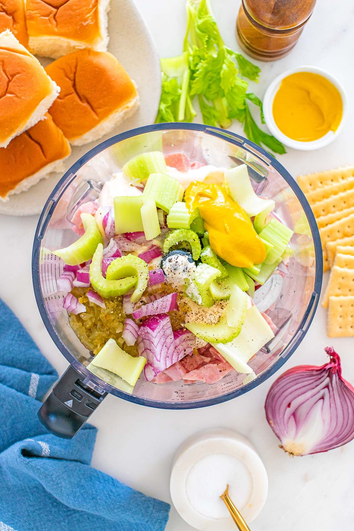 Chopped celery, red onion, and mustard are blended together in a food processor to create a classic ham salad, surrounded by rolls, crackers, and condiments.