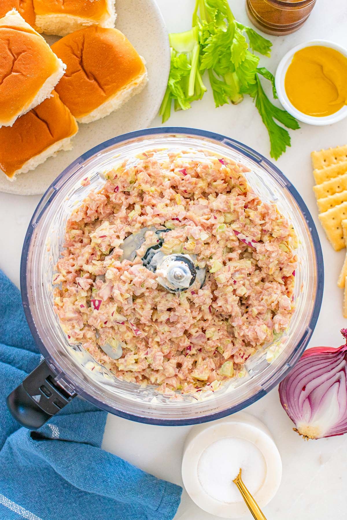 Chopped ham salad in a food processor, surrounded by buns, crackers, celery, mustard, and a red onion—a classic spread that showcases the savory flavor of ham salad.