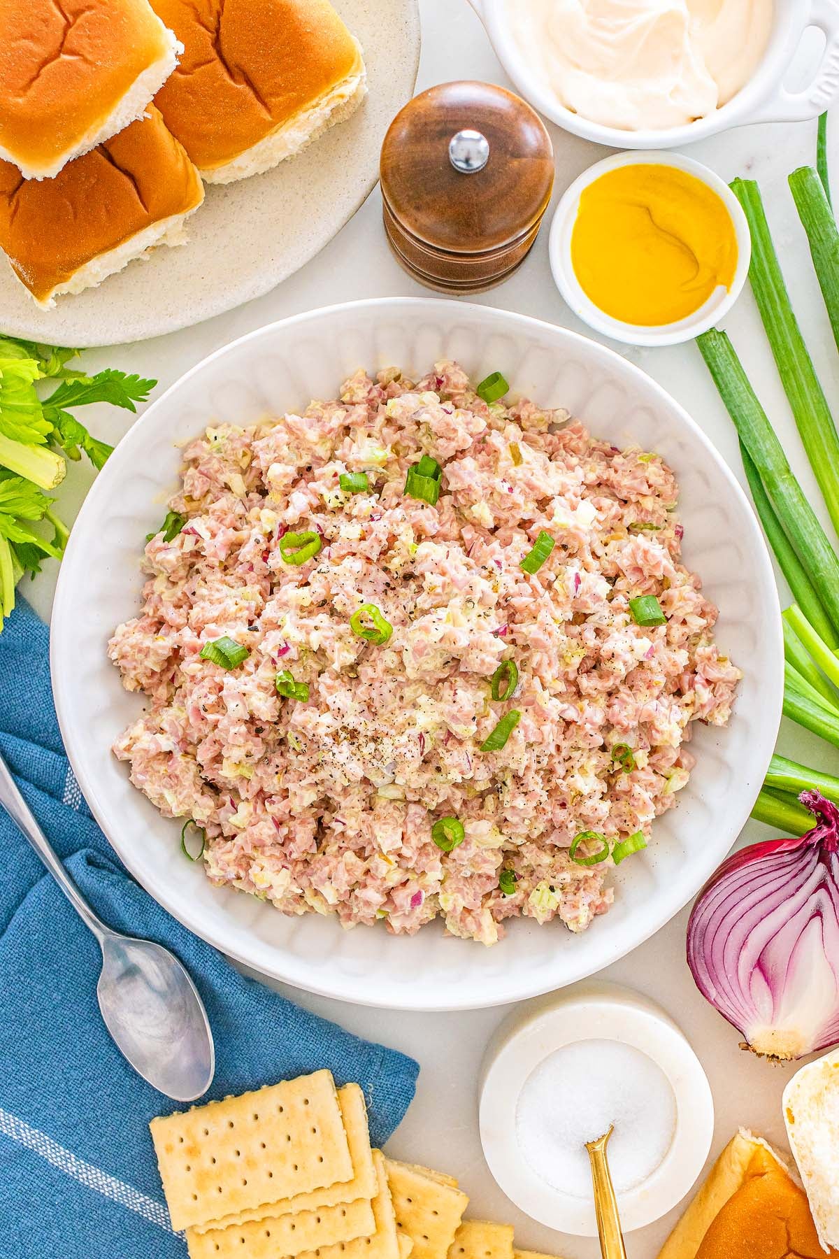 A bowl of ham salad garnished with green onions sits at the center, surrounded by crackers, rolls, and an assortment of condiments.