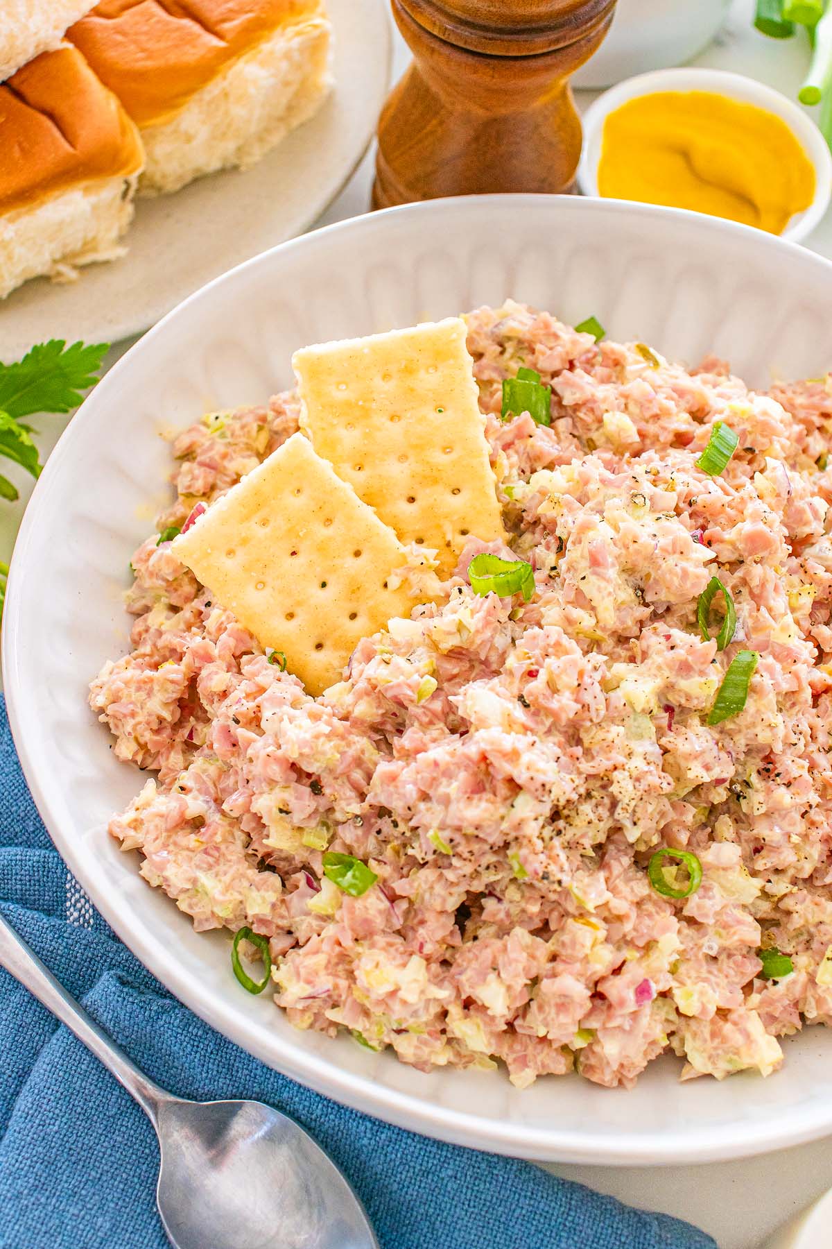 A bowl of ham salad topped with crackers and green onions, with rolls, mustard, and pepper in the background makes for a perfect savory snack or light meal.