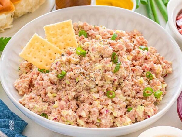 A bowl of creamy ham salad garnished with green onions and black pepper, served with two crisp saltine crackers.