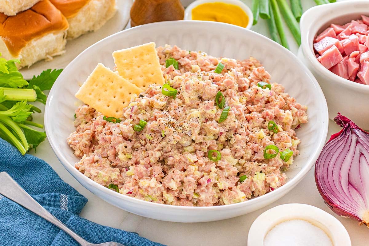A delicious bowl of ham salad with crackers on top, surrounded by fresh ingredients like onion, celery, and diced ham.