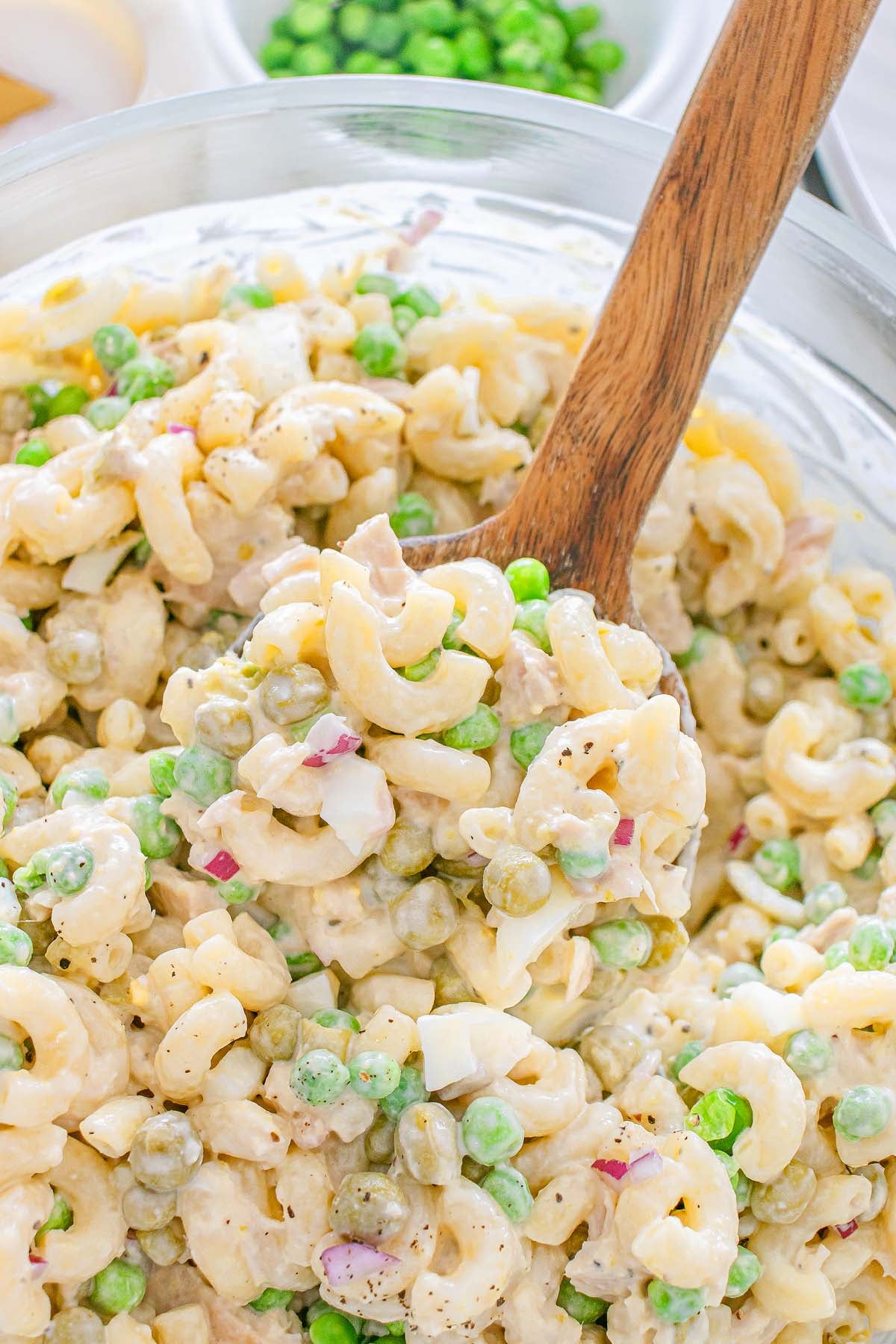 Spoon scooping creamy tuna macaroni salad with peas, eggs, and onions from a glass bowl.