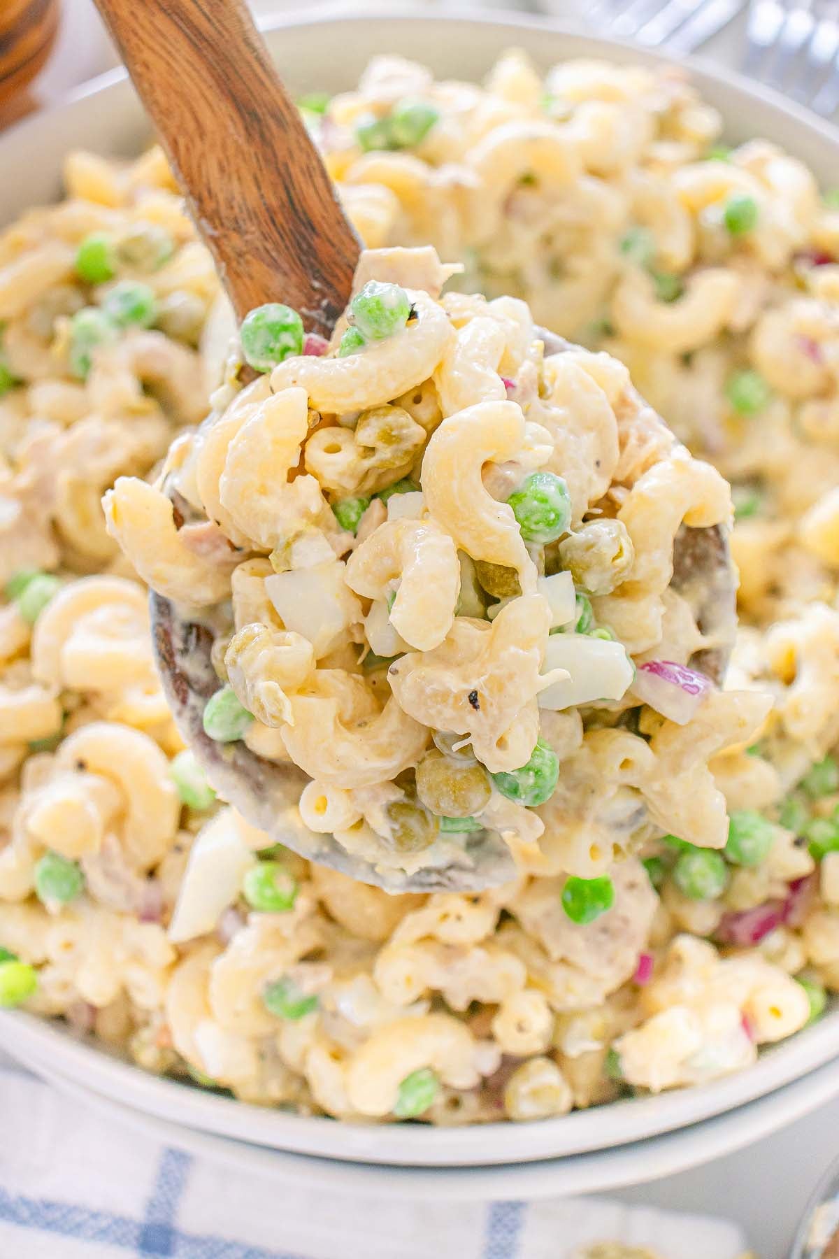 A spoonful of creamy tuna macaroni salad with green peas, onions, and elbow pasta above a bowl.