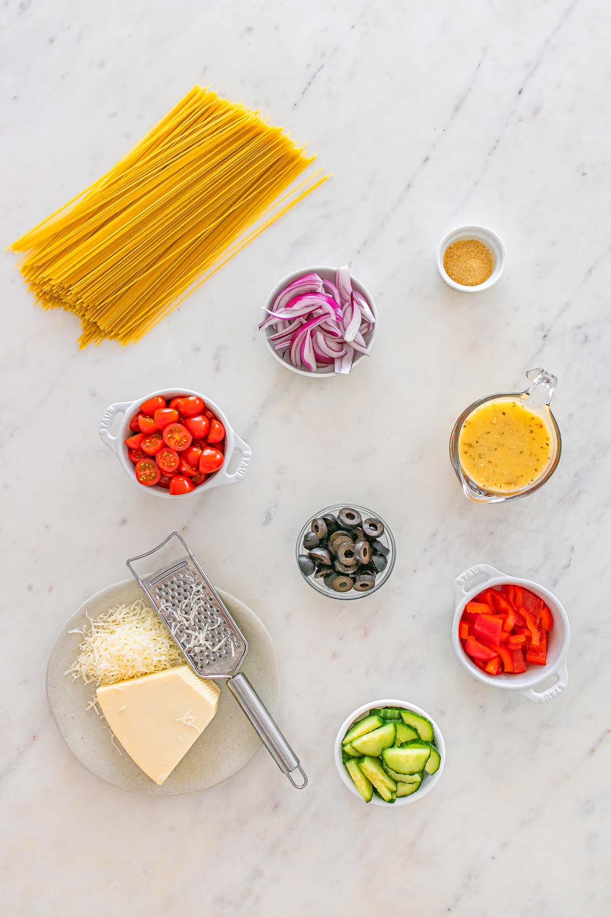 Uncooked spaghetti, veggies, olives, cheese, and dressing arranged on a marble surface.