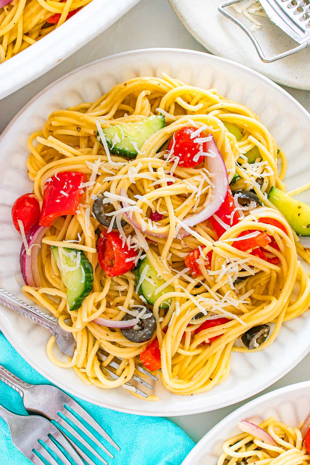 Spaghetti salad with tomatoes, cucumbers, olives, red onions, and shredded cheese on a white plate.