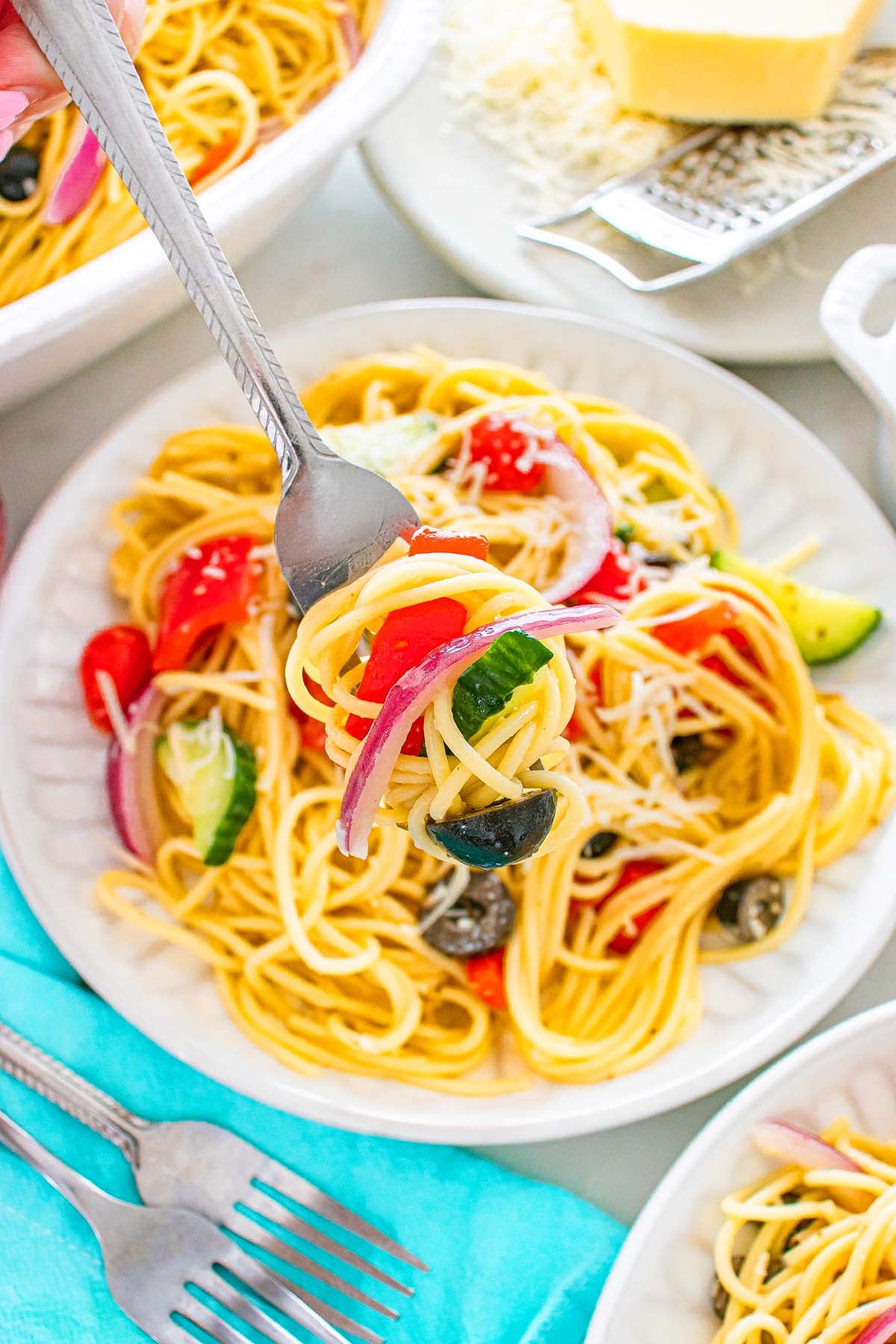 A fork lifts spaghetti with tomatoes, olives, zucchini, and red onion from a white plate.