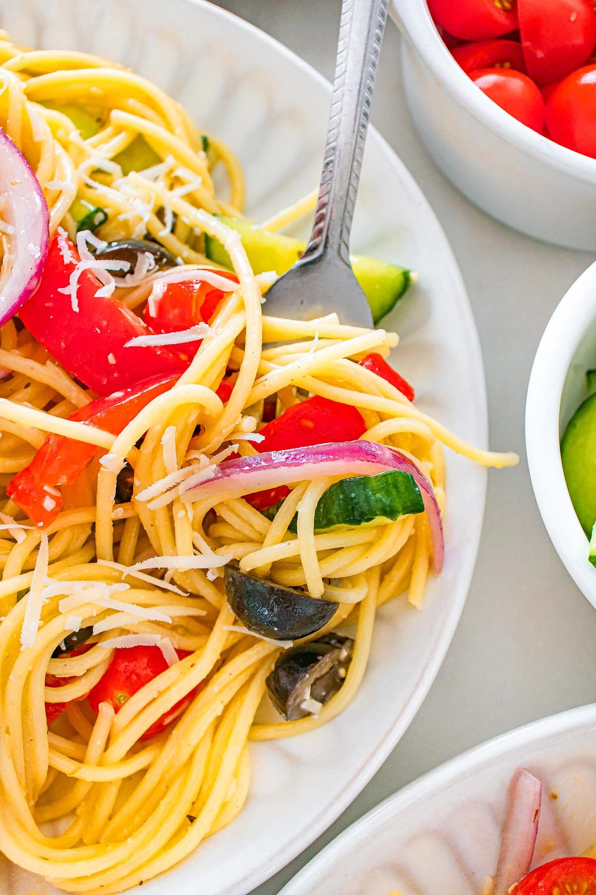 Plate of spaghetti salad with tomatoes, olives, cucumbers, red onion, and grated cheese, fork resting on plate.
