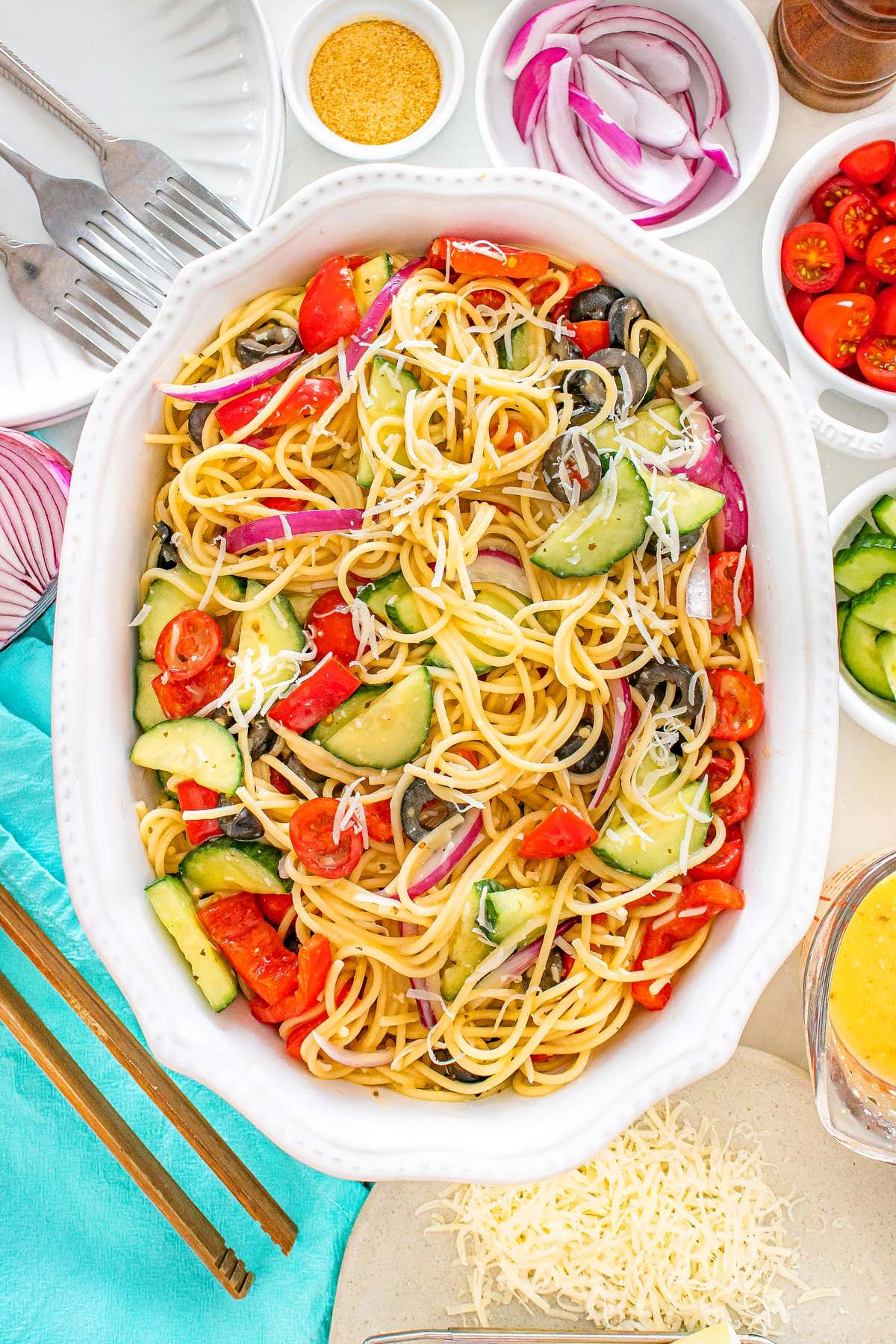 Pasta salad with tomatoes, cucumbers, olives, and red onions in a white dish, surrounded by toppings.