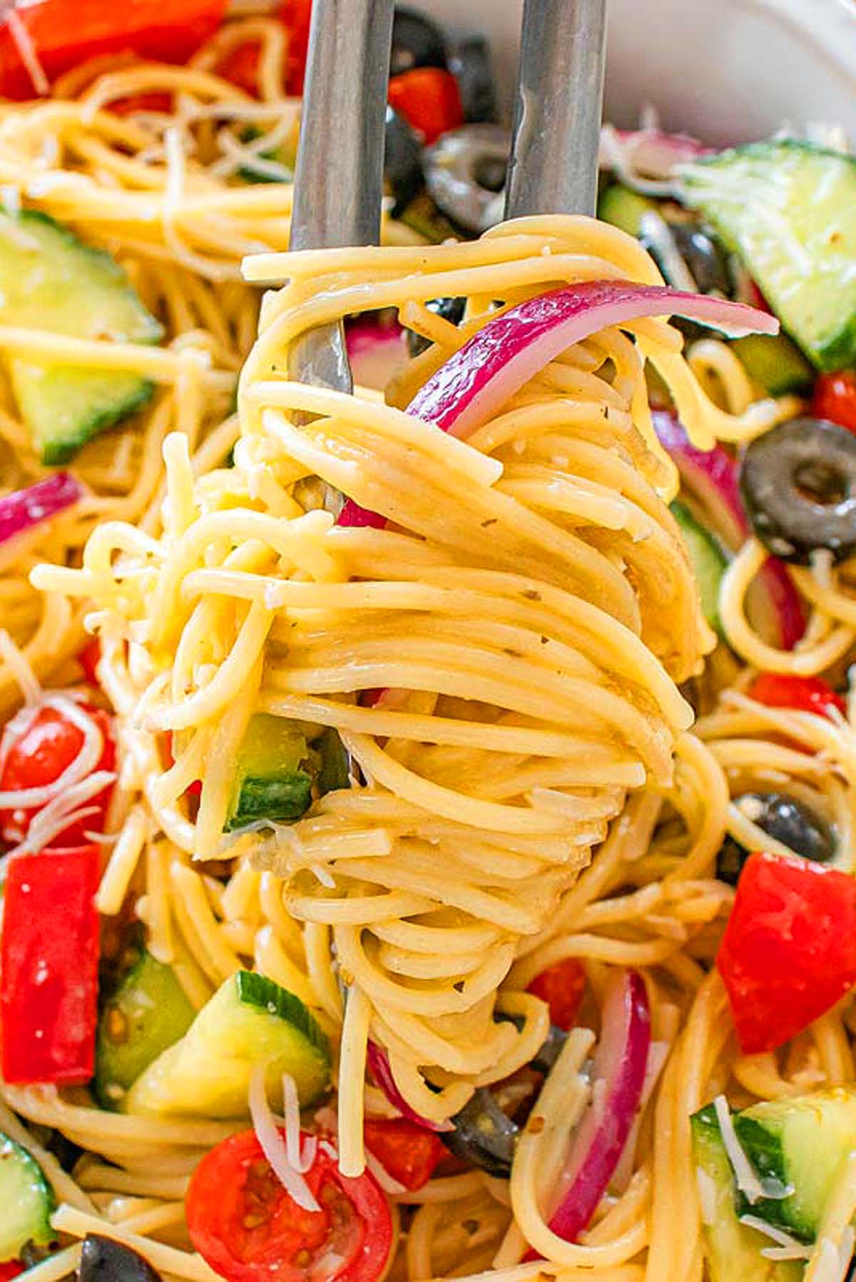 Tongs holding spaghetti with cucumbers, tomatoes, olives, red onion, and cheese in a pasta salad.