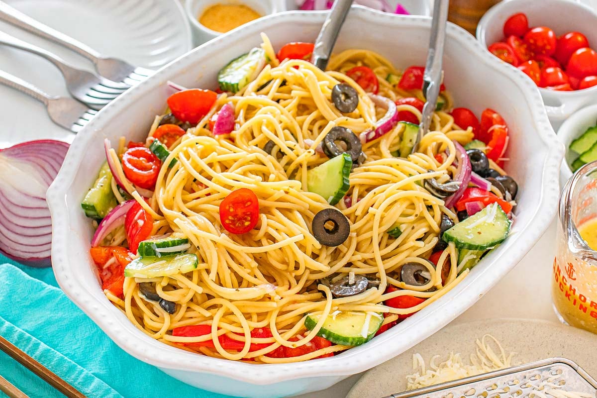 Pasta salad with tomatoes, cucumbers, olives, and onions in a white dish with serving tongs.