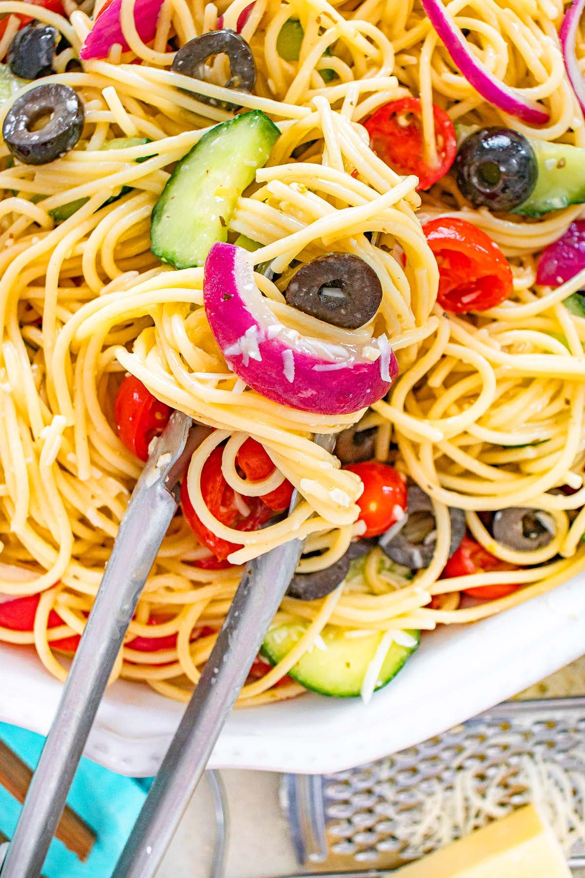 Tongs holding a serving of spaghetti pasta salad with olives, cherry tomatoes, cucumber, and red onions.