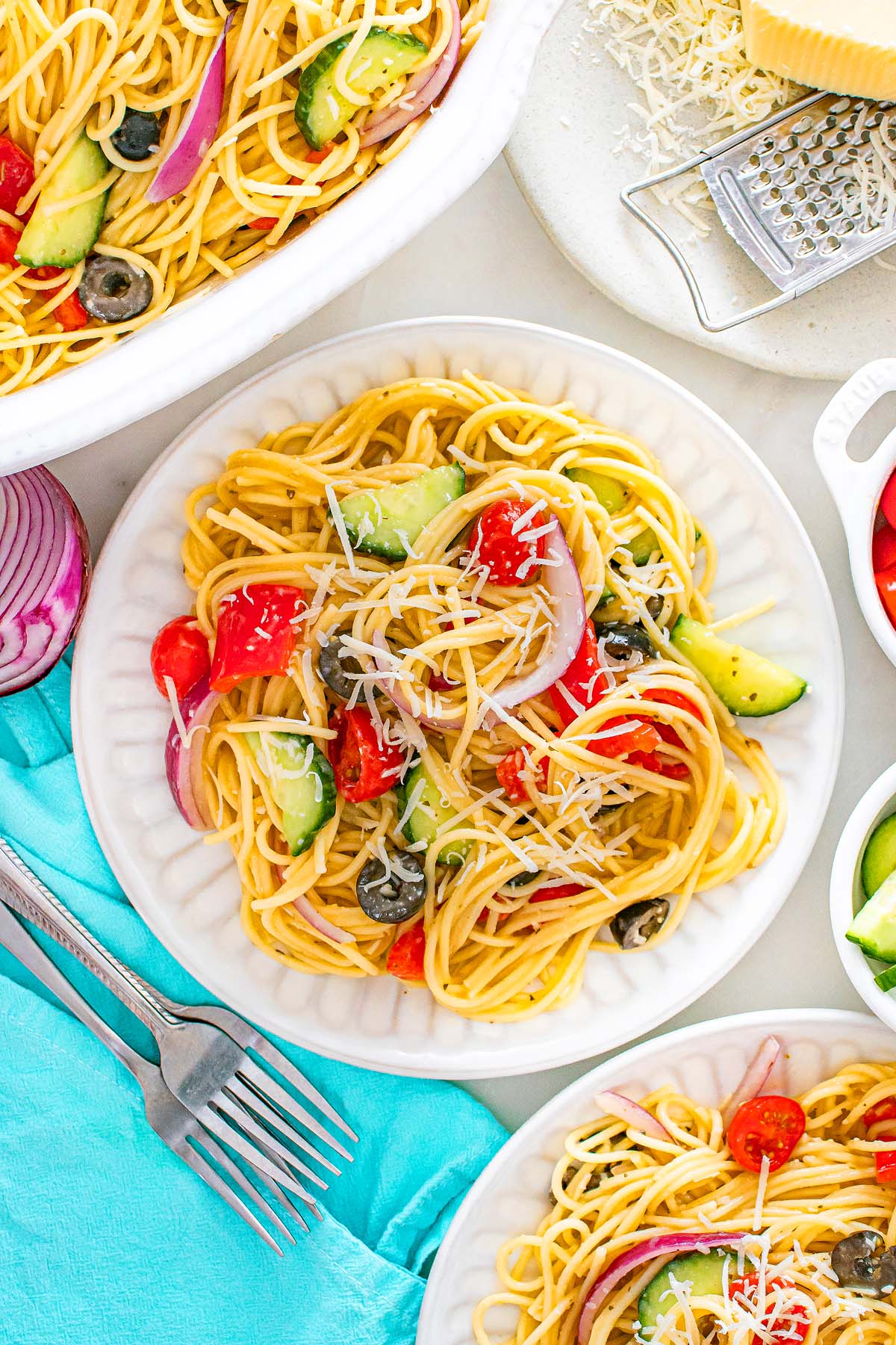 Spaghetti pasta salad with tomatoes, cucumbers, olives, onions, and shredded cheese on white plates.