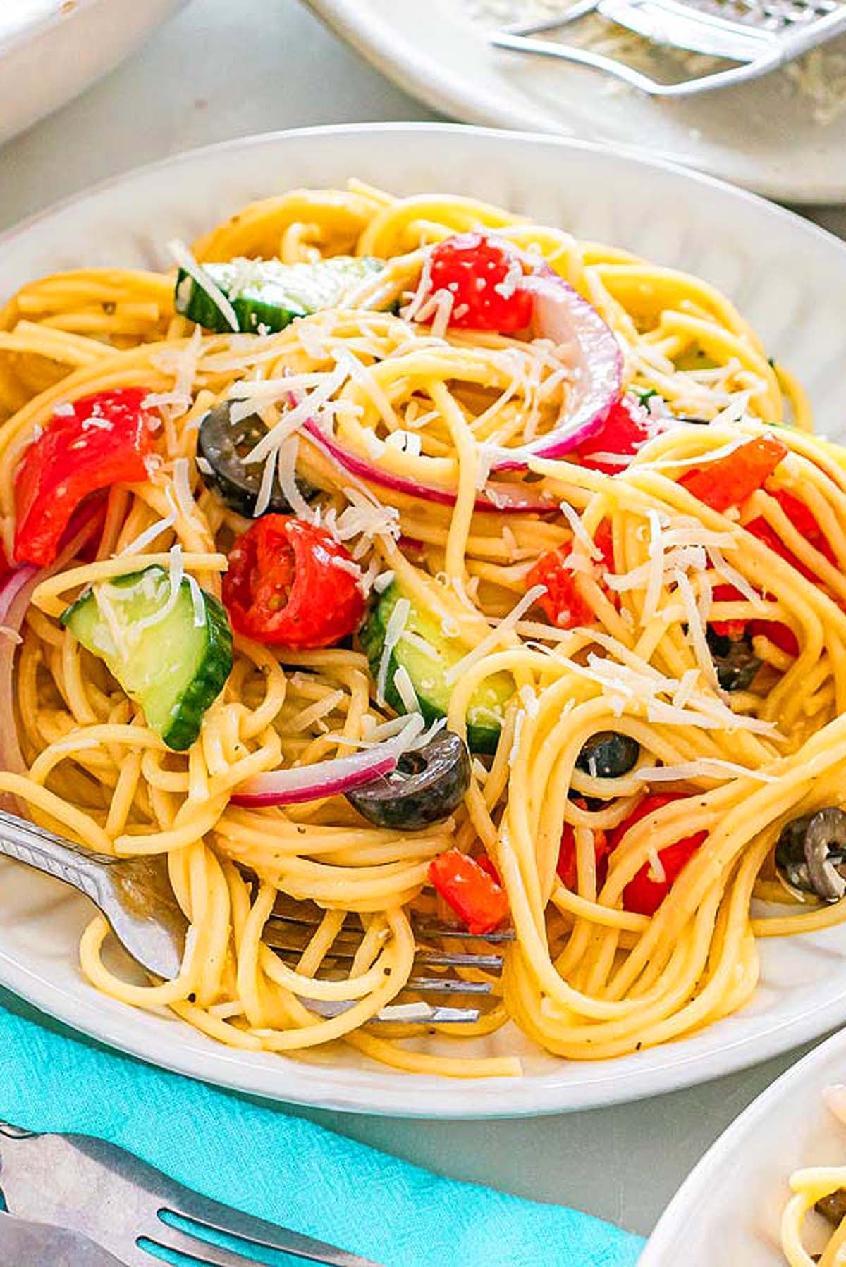 Spaghetti salad with tomatoes, cucumbers, olives, red onions, and shredded cheese on a white plate.