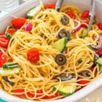 Pasta salad with cherry tomatoes, cucumbers, olives, and red onions in a white serving dish with tongs.