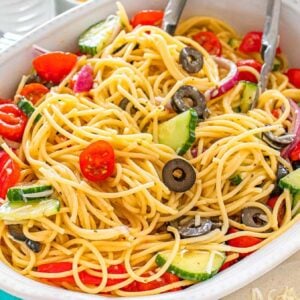 Pasta salad with cherry tomatoes, cucumbers, olives, and red onions in a white serving dish with tongs.