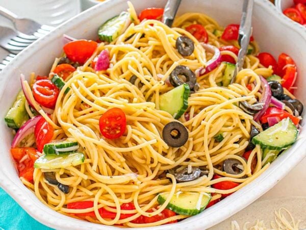 Pasta salad with cherry tomatoes, cucumbers, olives, and red onions in a white serving dish with tongs.