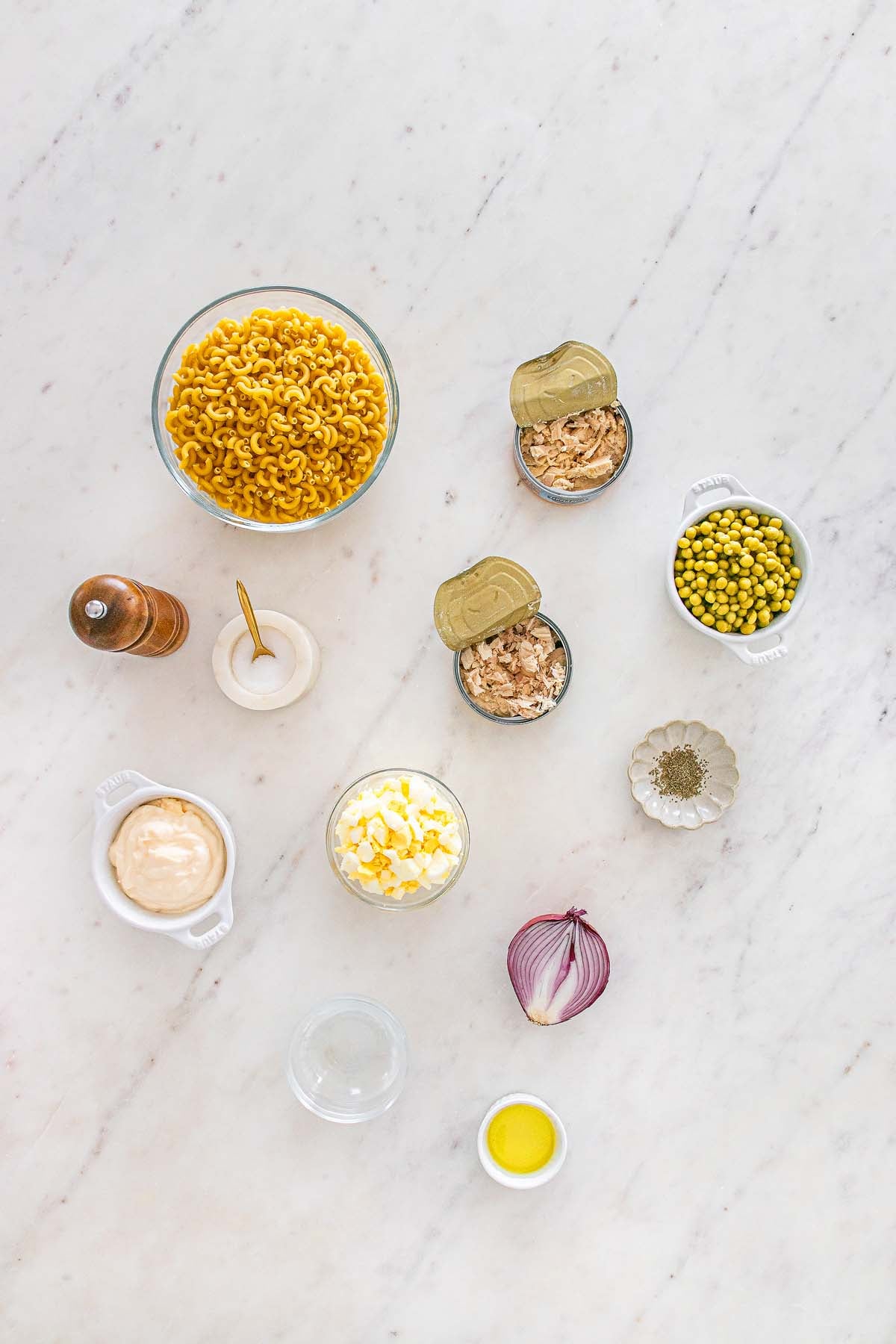 Ingredients for a classic tuna macaroni salad, including pasta, tuna, peas, onion, mayo, eggs, and seasonings arranged on a marble surface.