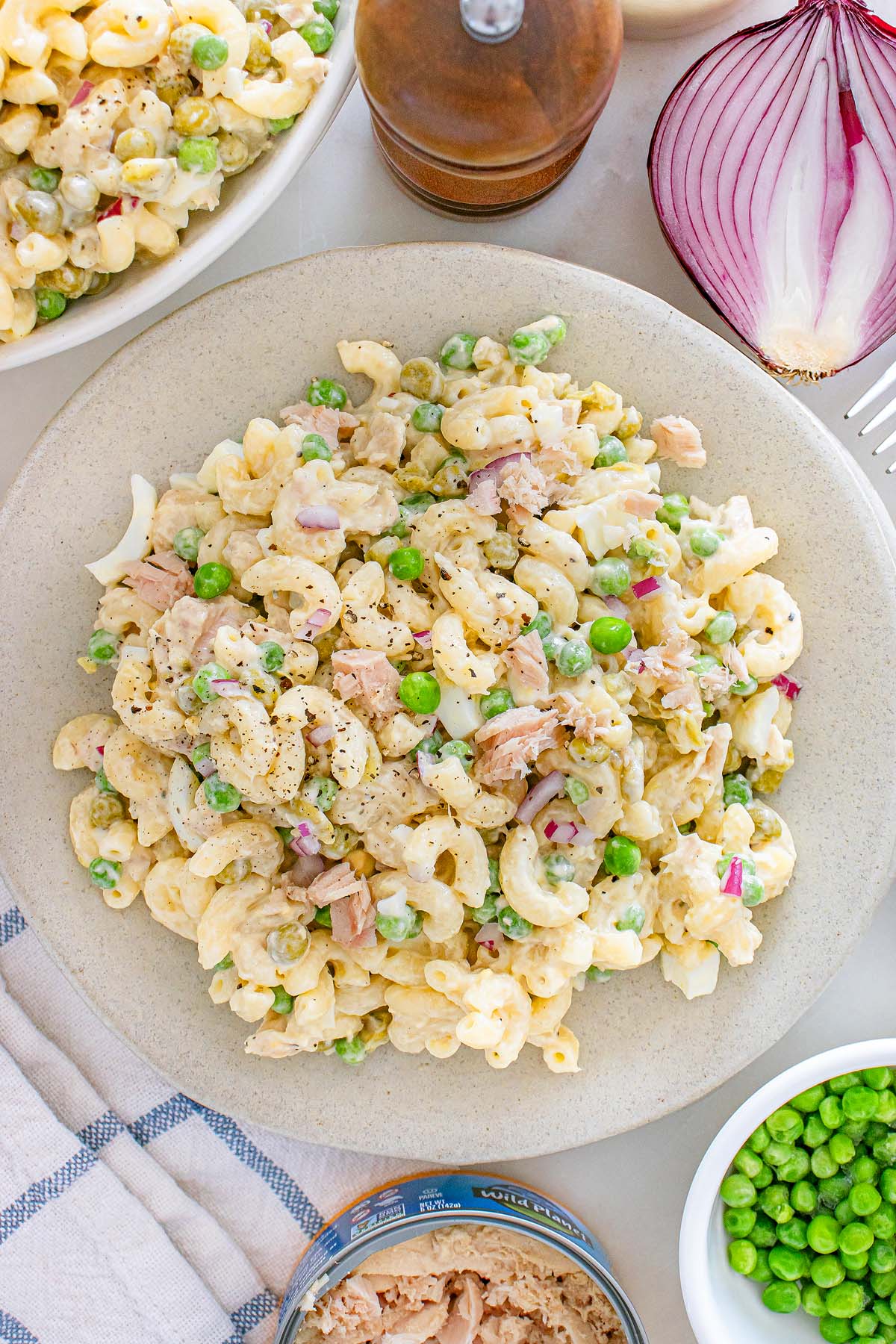 Plate of tuna macaroni salad with peas, red onion, and black pepper, surrounded by fresh salad ingredients for a classic and flavorful tuna macaroni salad experience.