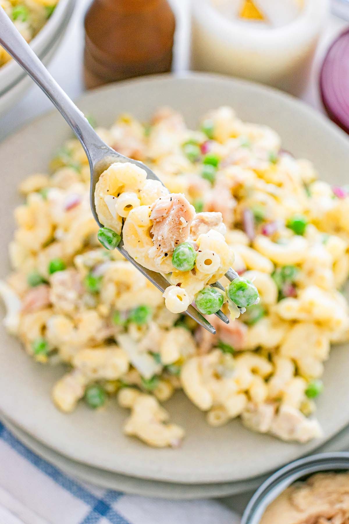 A fork holds a bite of macaroni salad with peas and ham over a plate filled with more of the classic tuna macaroni salad, combining savory flavors in every bite.