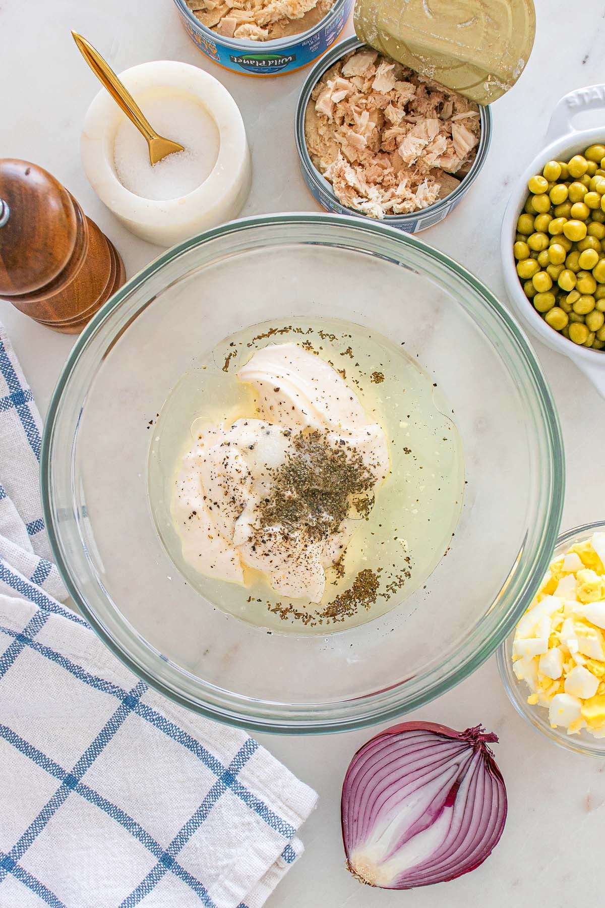 Glass bowl with mayo, spices, onions, and eggs; tuna, peas, salt, pepper, and a towel around it—everything you need to prepare a classic tuna macaroni salad.