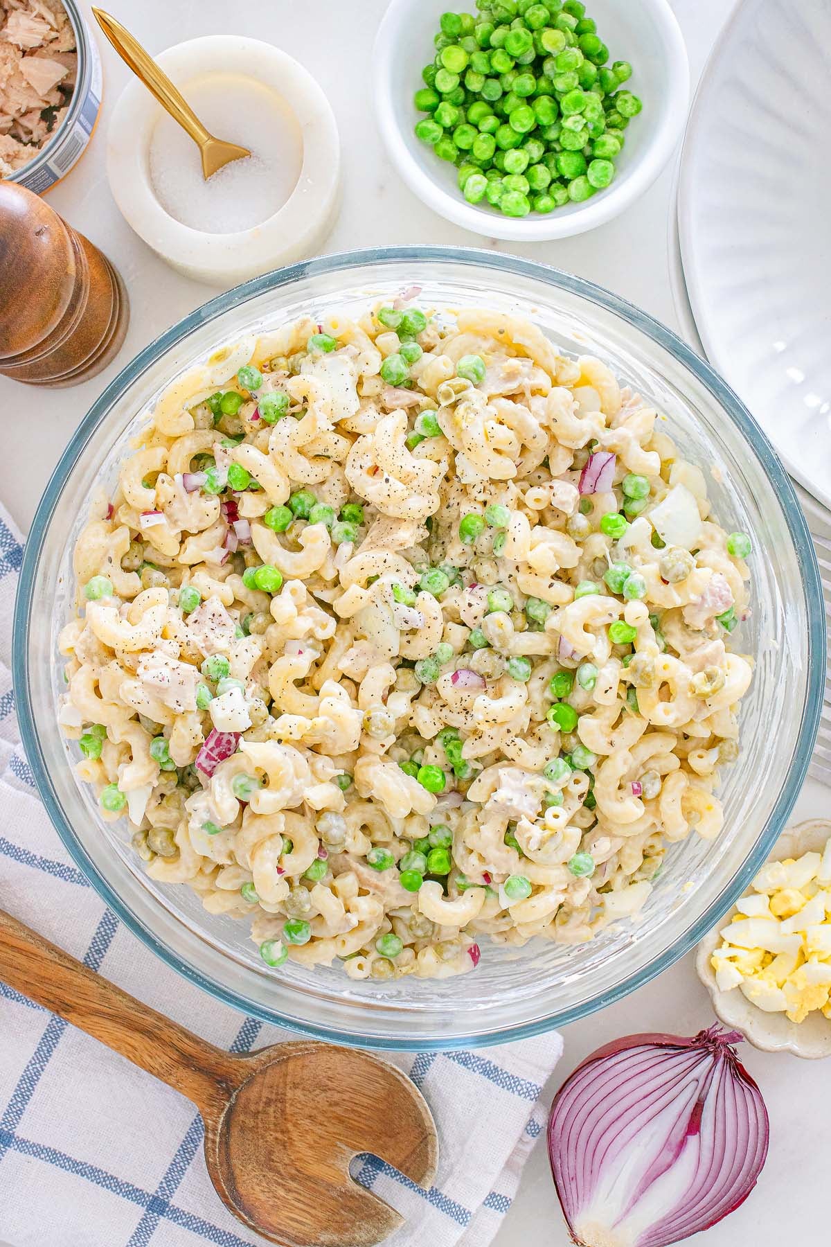 A bowl of tuna macaroni salad with peas and red onion sits on a table, surrounded by fresh ingredients and utensils nearby.