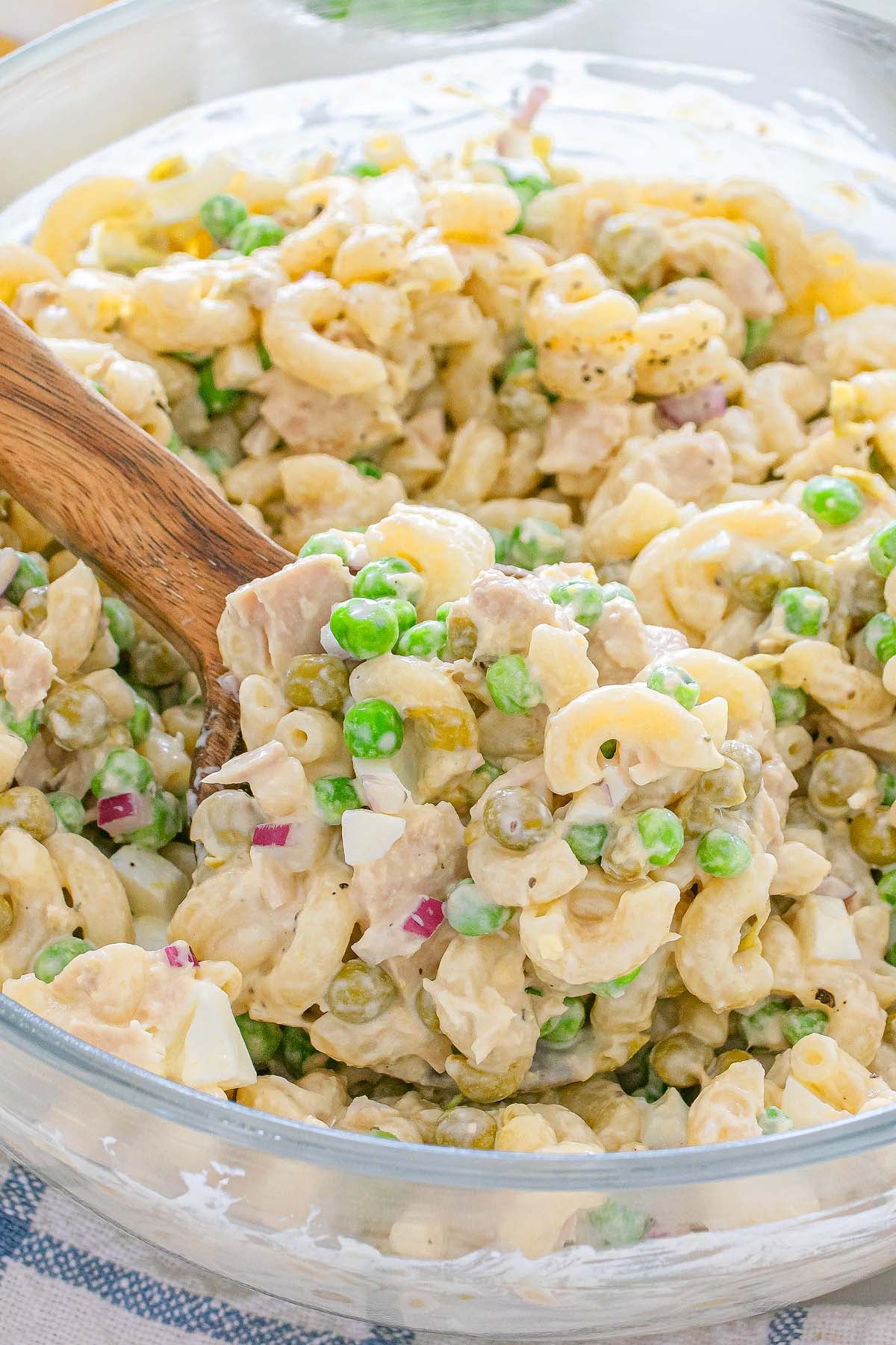 Tuna macaroni salad with peas, diced onions, and a creamy dressing served in a glass bowl with a wooden spoon.