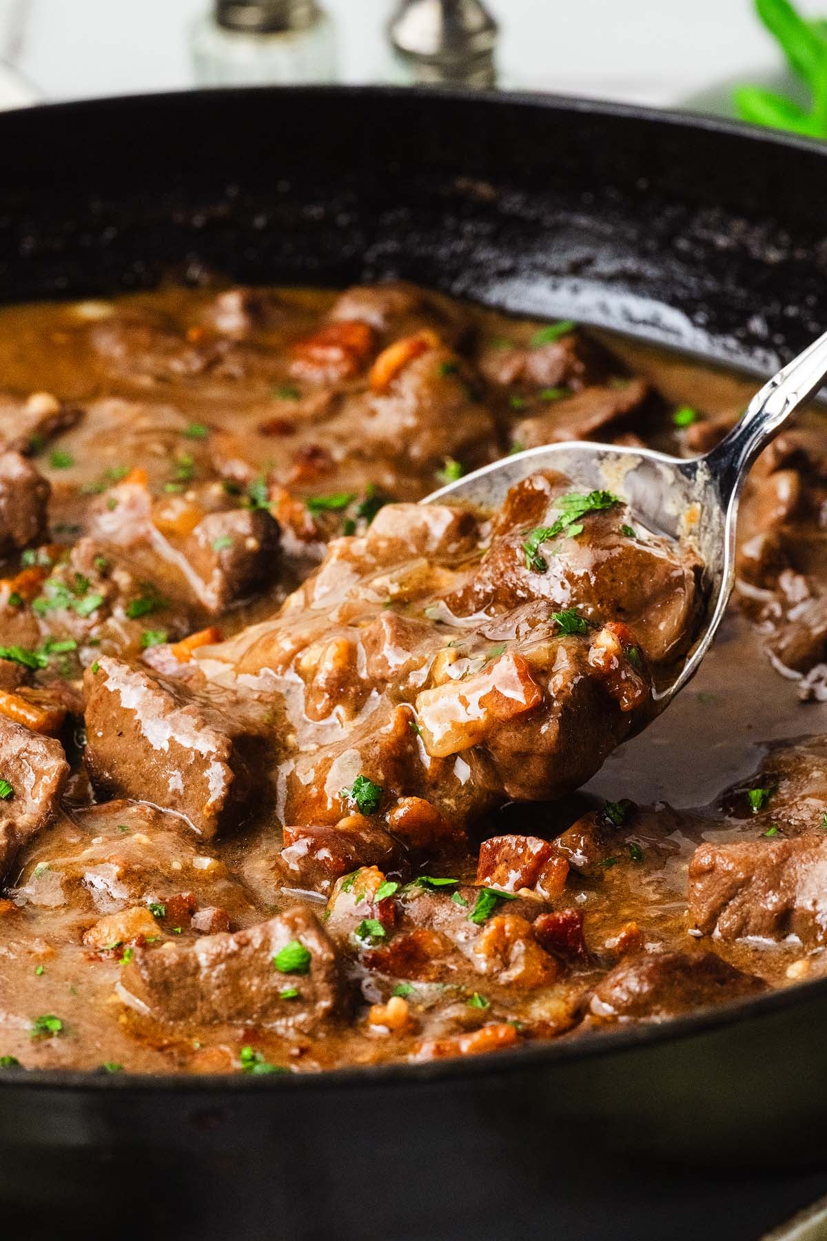 Spoon serving beef tips with gravy, carrots, and herbs from a black pan.
