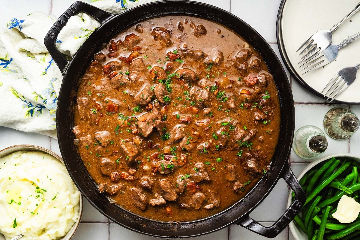 A skillet of beef tips and gravy with mashed potatoes, green beans, and plates with forks on the side.