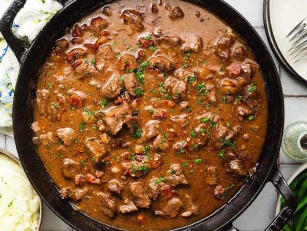 Beef tips and gravey in a black skillet, served with mashed potatoes, green beans, and plates with forks on the side.